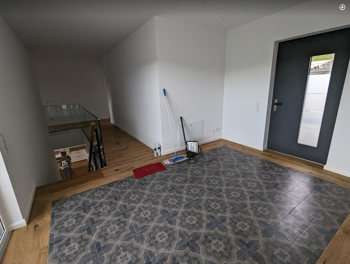 Heller Eingangsbereich mit dunkelgrauer Tür, gemustertem Fliesenboden und Reinigungswerkzeug.