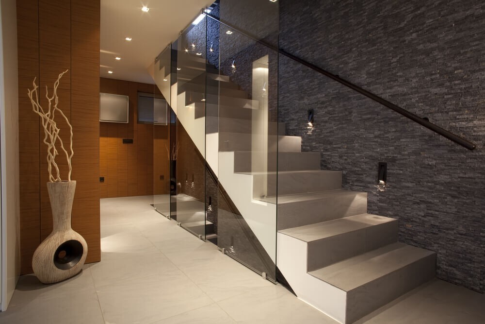 Moderner Innenraum mit weißer Treppe, Glasgeländer, dunkler Steinwand und Vase mit Zweigen.