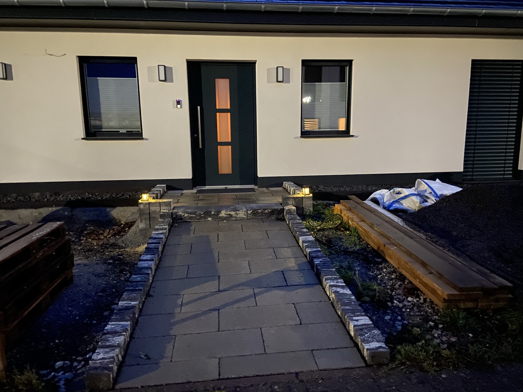 Nachtaufnahme moderner Hausfront: dunkle Tür, Fensterpaar, beleuchteter Steinweg, Holzbohlen rechts.