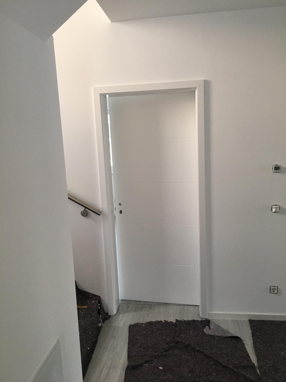 Weißer Innenflur mit geschlossener Tür, linkem Treppenhandlauf und dunklen Teppichen.