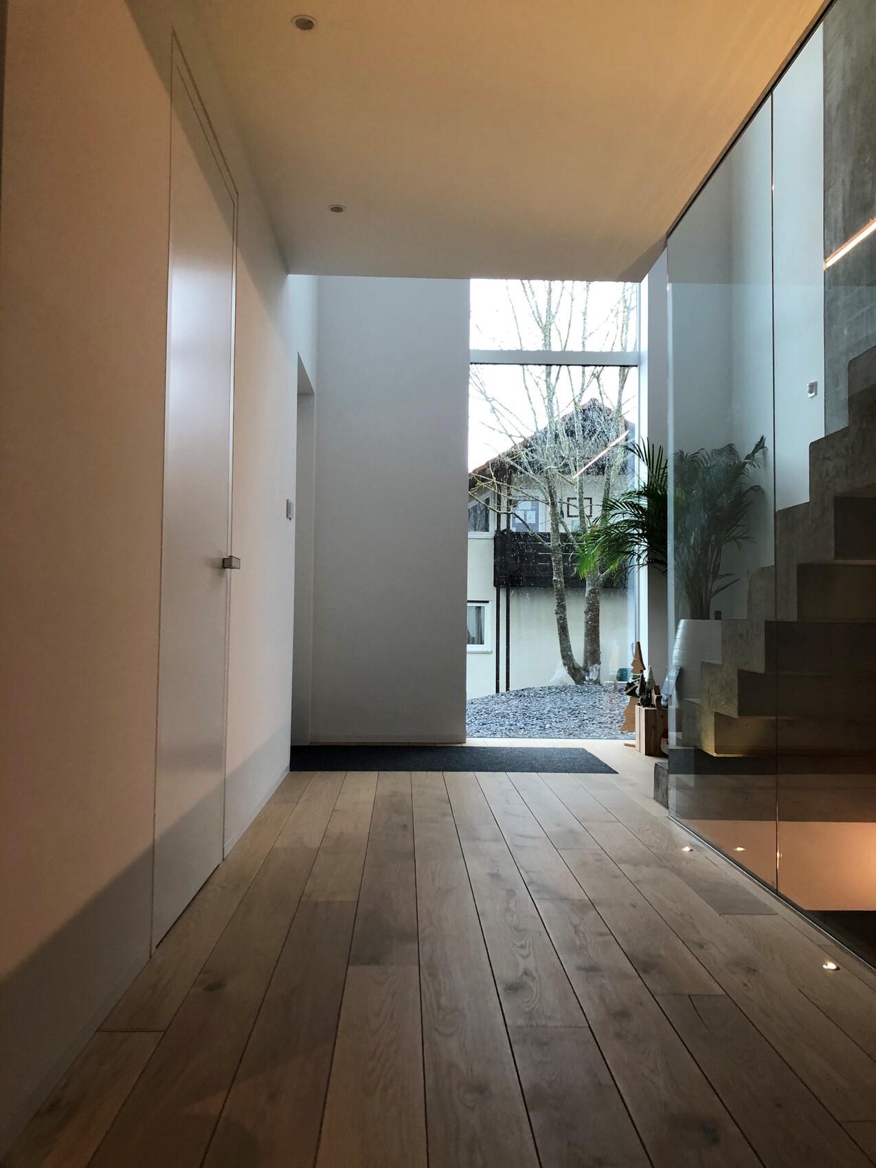 Moderner Flur mit Holzfußboden, Glaswand rechts, Treppe und Sicht auf Garten außen.