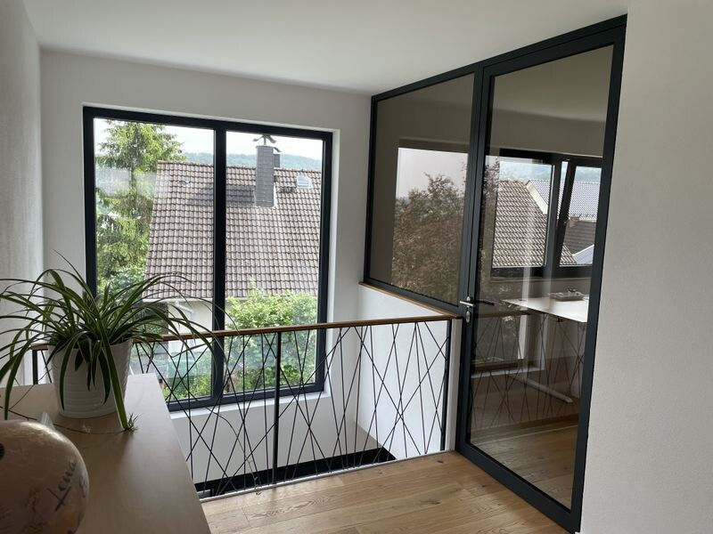 Heller Innenraum mit großer Fensterfront, Blick auf Dächer, Glastür, Geländer und Pflanze.
