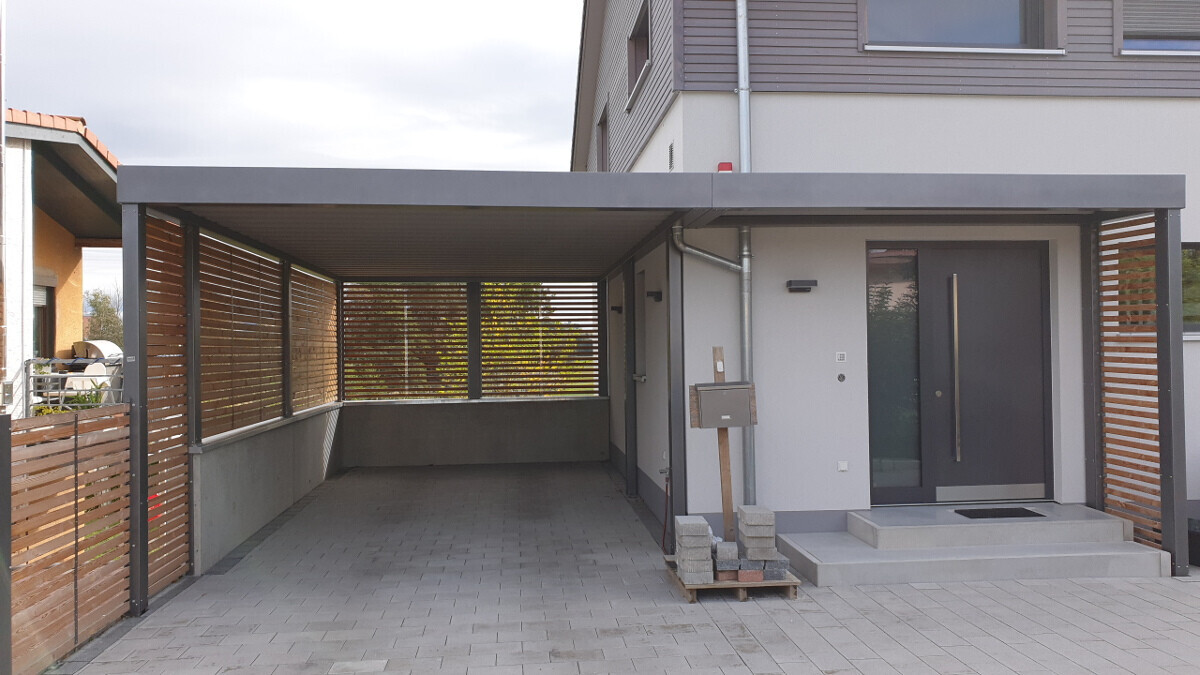 Moderne Hauszufahrt mit Carport, seitlichen Holzlamellen, grauer Eingangstür und Briefkasten.