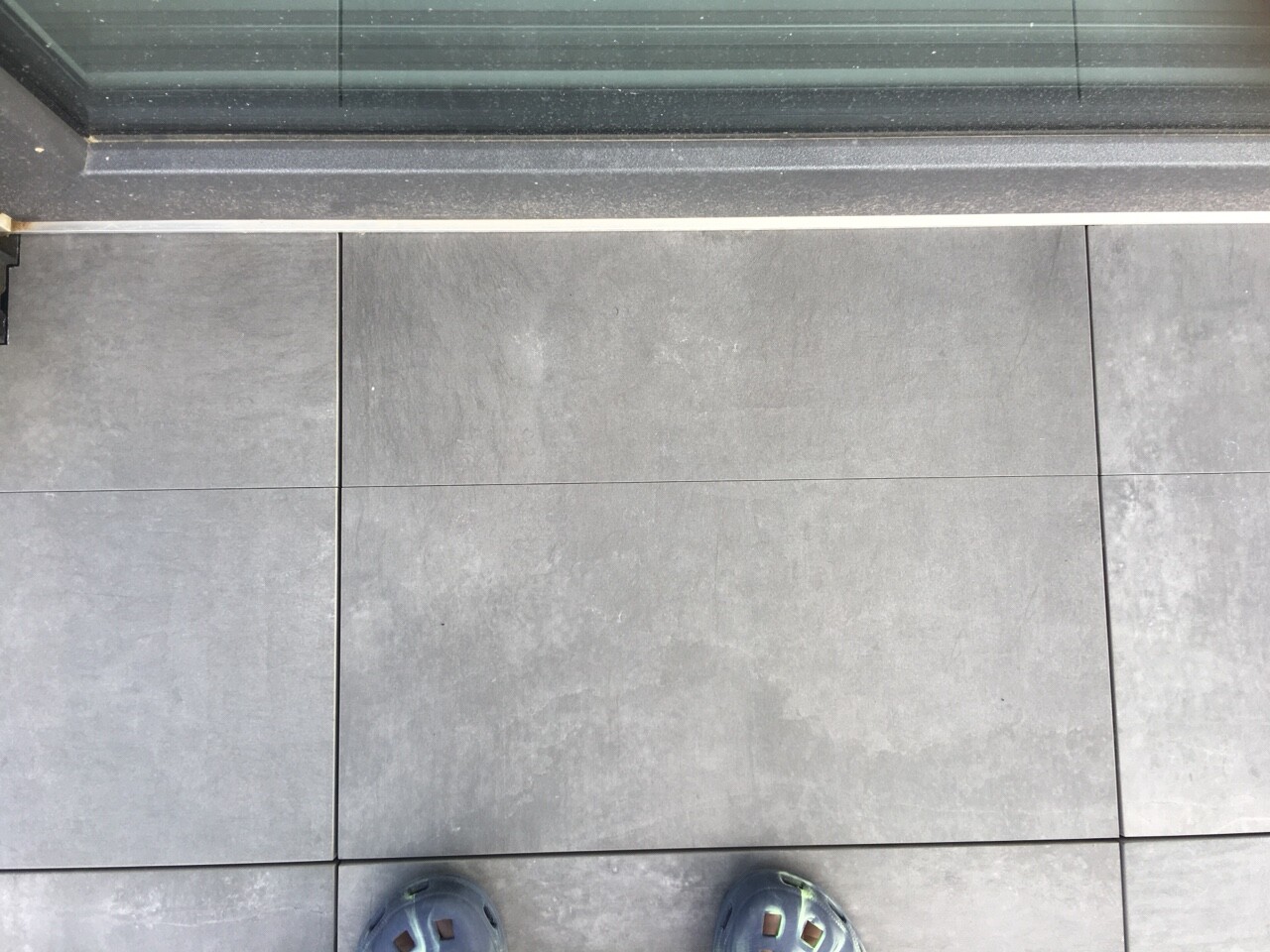 Blick auf graue Bodenfliesen mit Fugen; obere Metall-Schwelle; zwei blaue Schuhe unten im Bild.