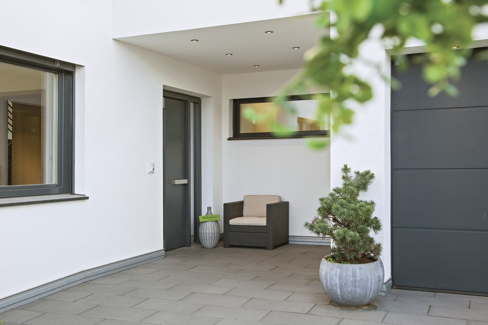 Eingangsbereich eines modernen weißen Hauses mit schwarzer Tür, Korbsessel und Pflanzkübel.