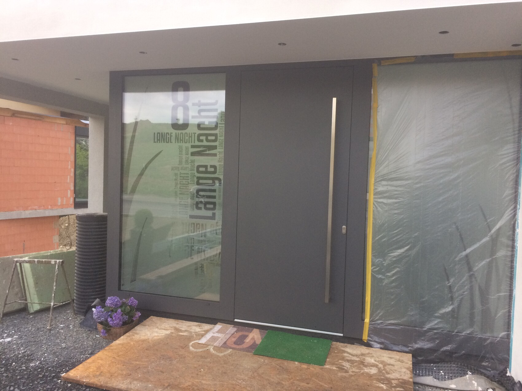 Moderne graue Haustür mit langem Griff; links Glasfenster, rechts verpacktes Fenster, Baubereich.