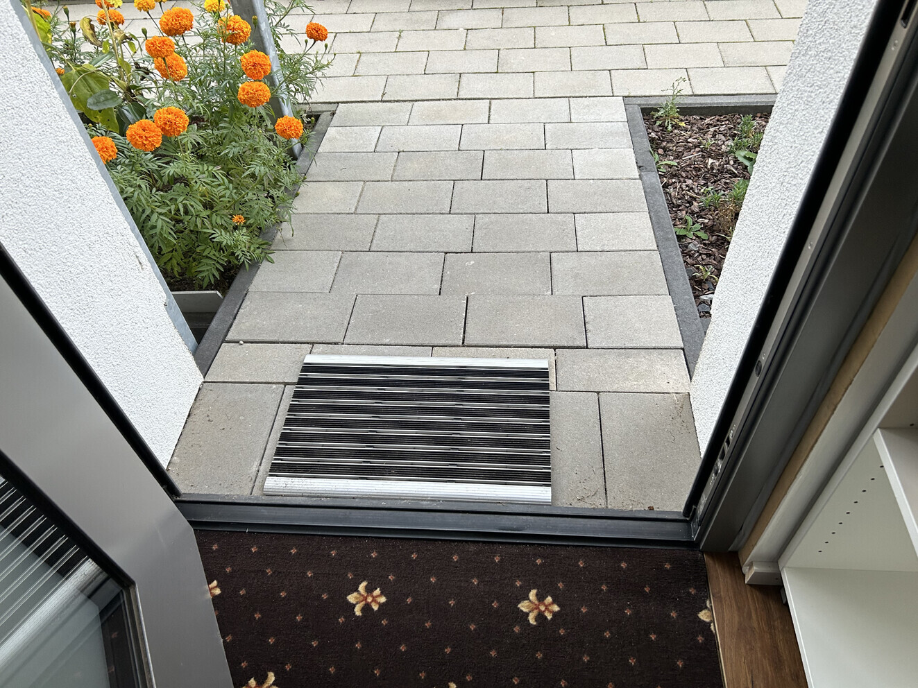 Offene Tür: gepflasterter Eingang, links orange Ringelblumen, rechts Beet, Gitter vor der Tür.