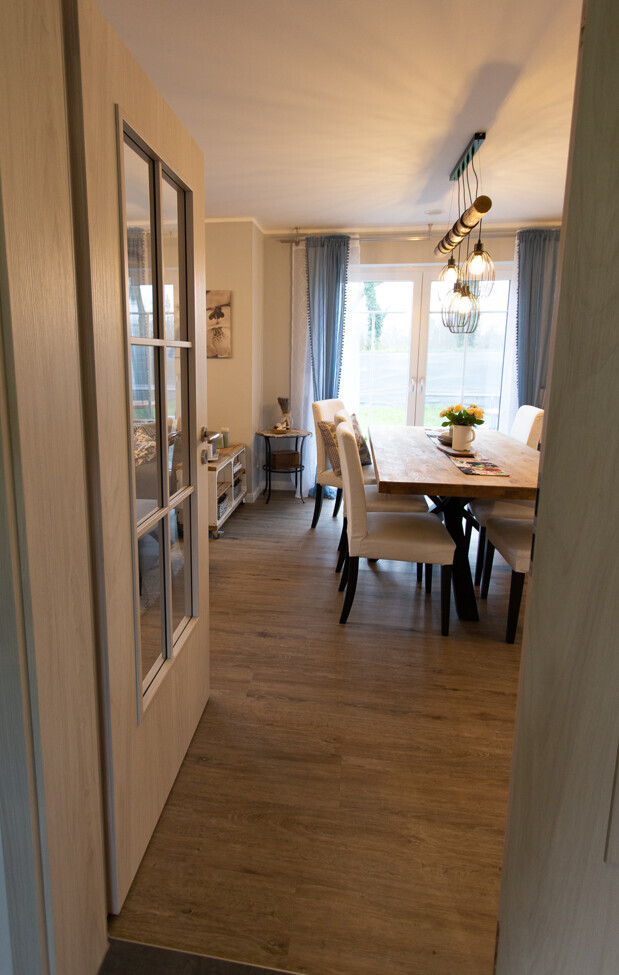 Helles Esszimmer mit Holztisch, gepolsterten Stuehlen, Kronleuchter, Vorhaengen und grossen Fenstern.