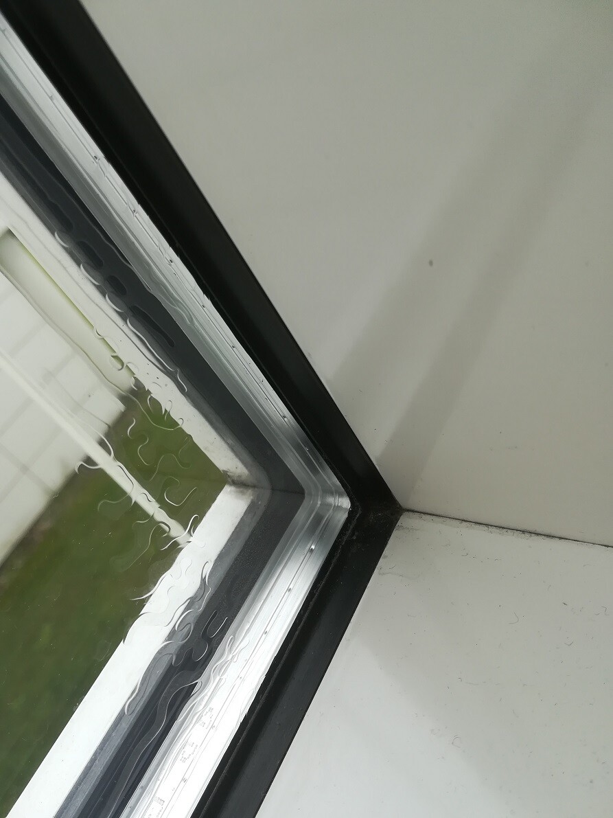 Fensterecke mit schwarzem Rahmen, Regen am Glas und weißer Wand; Blick nach draußen auf Grünfläche.