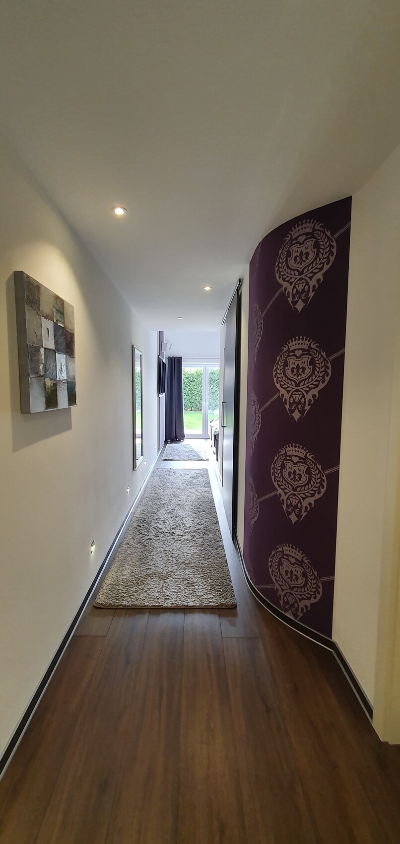 Langer Flur mit beigem Teppich, linke Kunst, rechte violette Wand mit Mustern, Tür zum Garten.