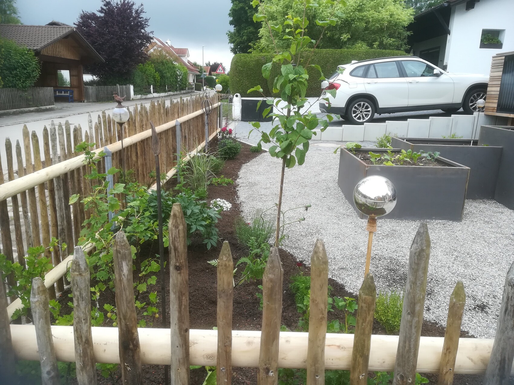 Gartenweg hinter Holzzaun mit Beetpflanzen, Baum, Kugellampe und weißem SUV im Hintergrund.