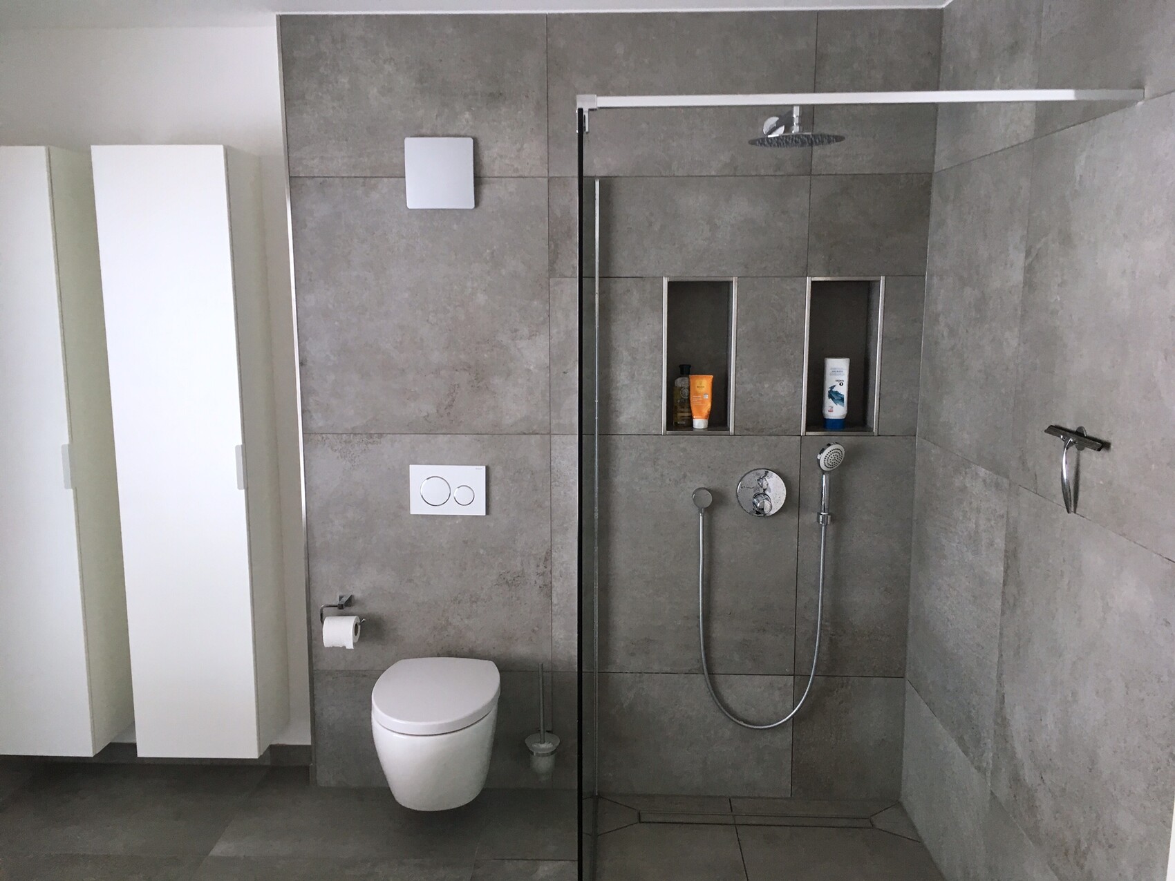 Modernes Badezimmer mit grauen Fliesen, wandhängendes WC, Glasdusche und Ablagen.