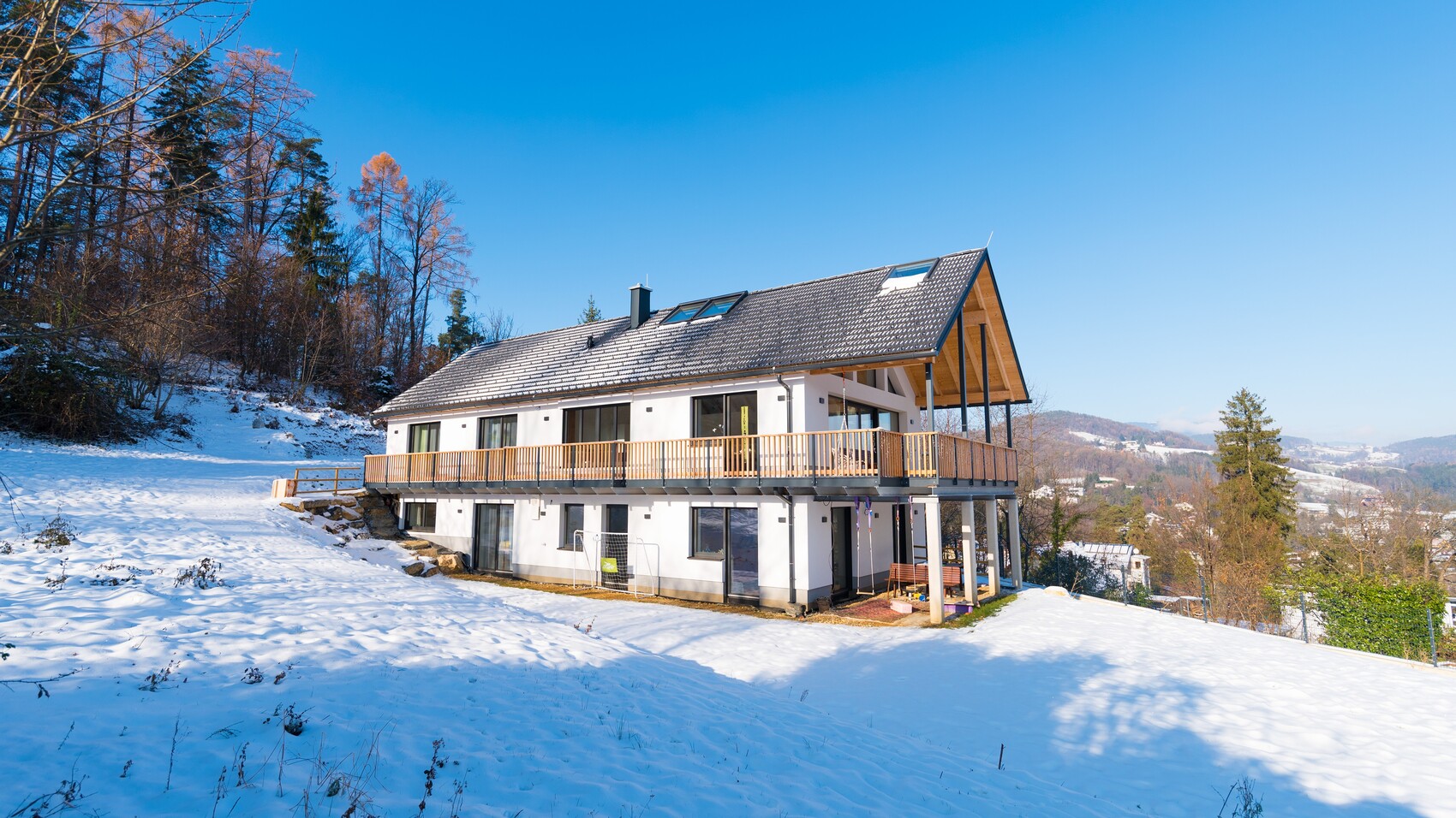 Modernes Haus mit großer Holzveranda, verschneitem Hang, Wald im Hintergrund, blauer Himmel.