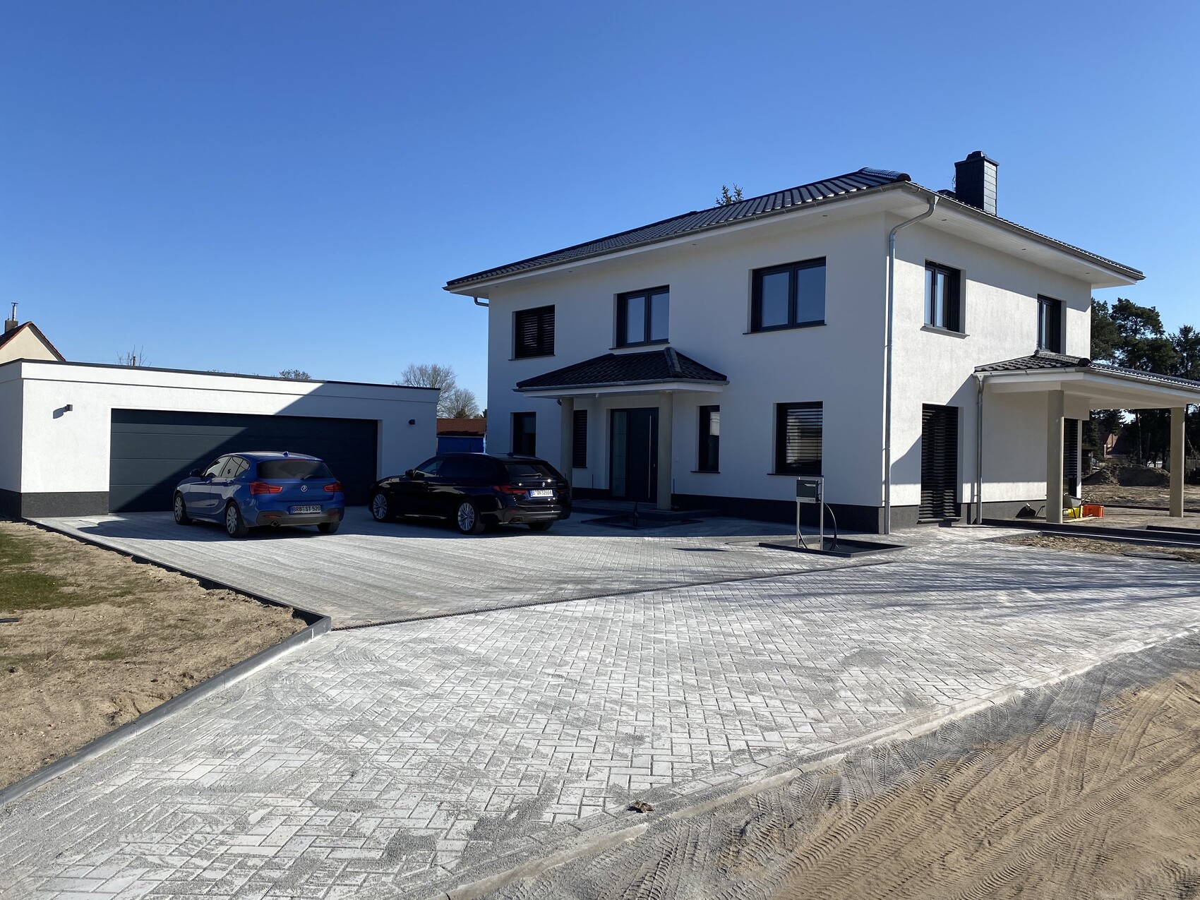 Modernes weißes Einfamilienhaus mit Garage links, zwei Autos auf gepflasterter Einfahrt.