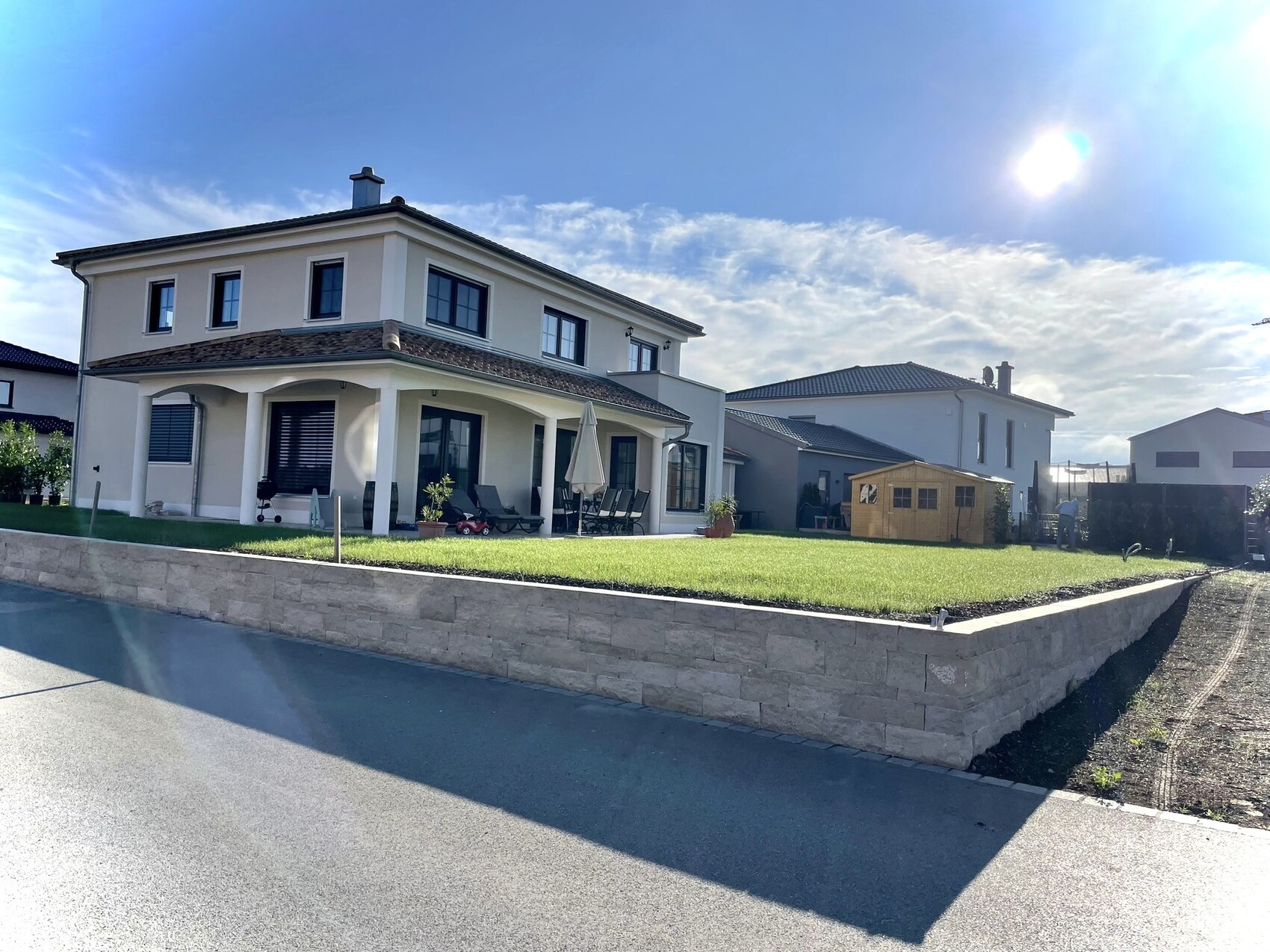 Zweistöckiges helles Haus mit Terrasse, Gartenmöbeln, Sonnenschein und Holzschuppen.