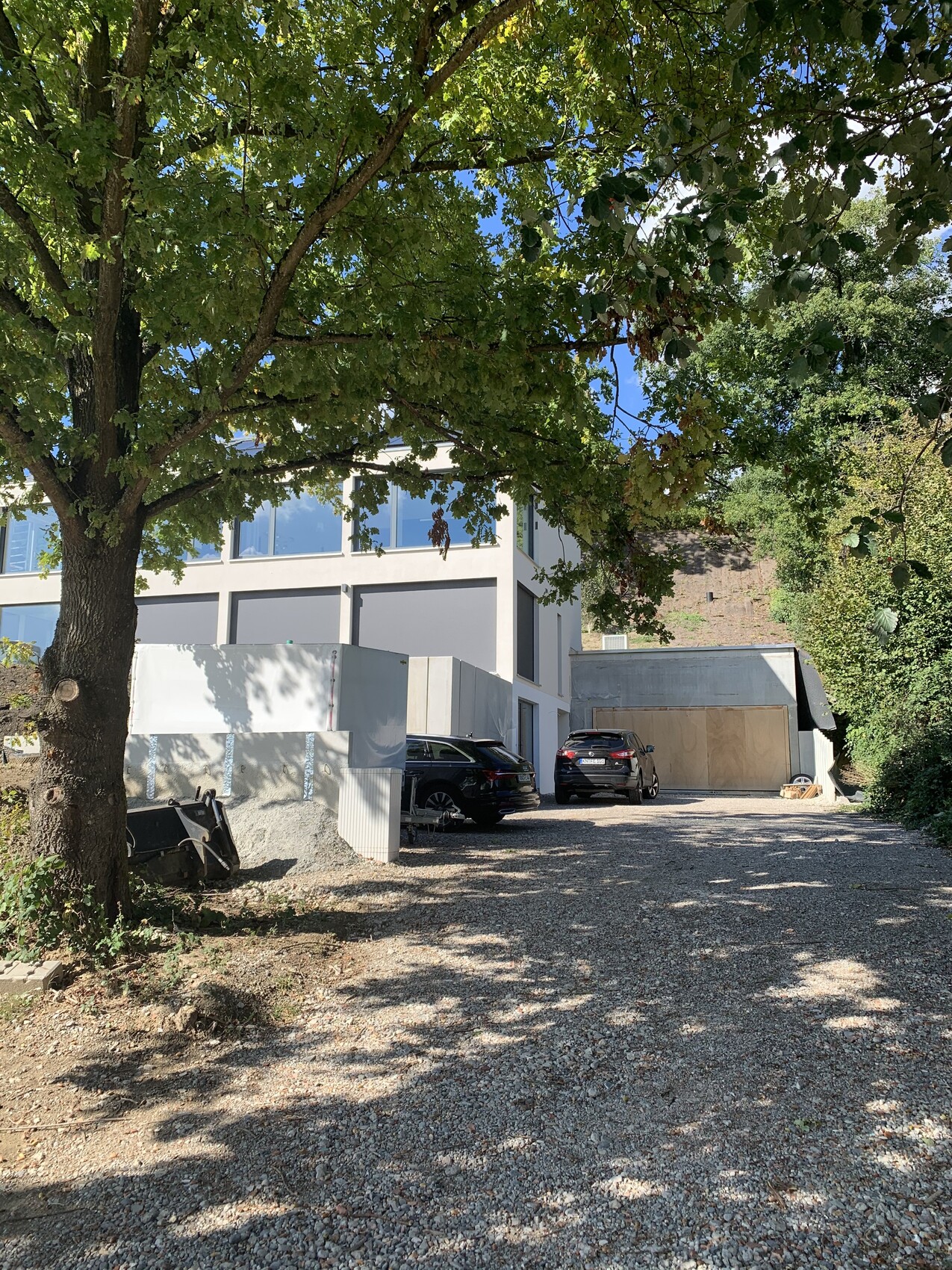 Großer Baum links, modernes weißes Gebäude mit Glasfenstern, Kiesweg und geparkten Autos.