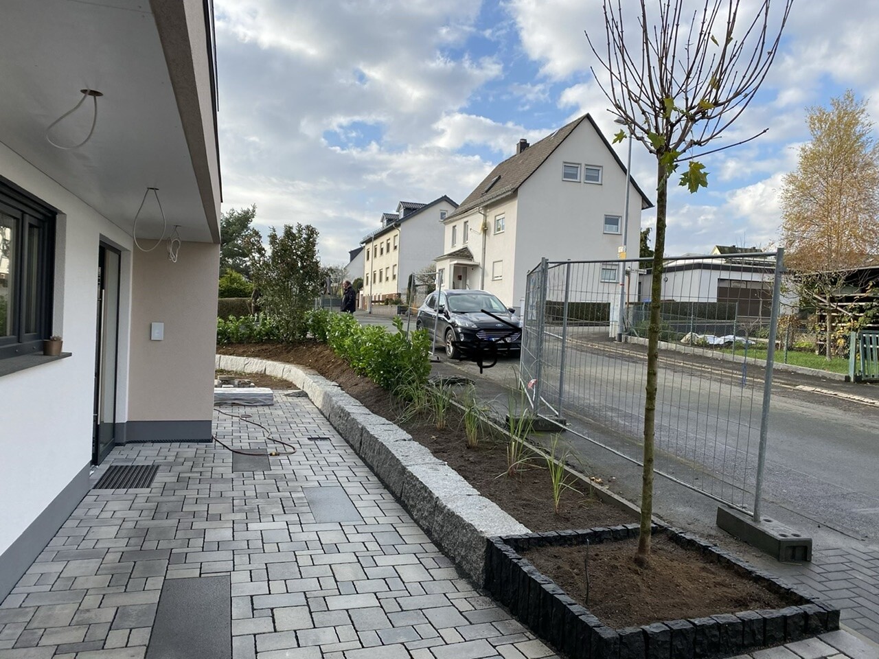 Gepflasterter Gehweg, Bauzaun, Baum im Beet und geparkter Wagen vor Häusern.