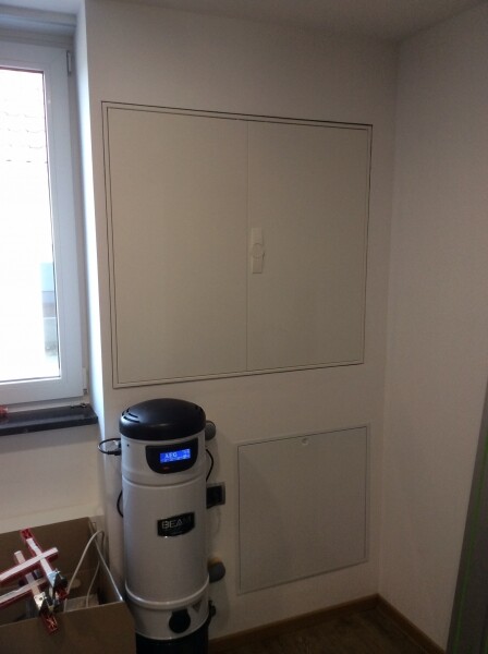 Kleine Innenraum-Ecke mit weißem Wandschrank, Zentralstaubsauger-Tank und Fenster.