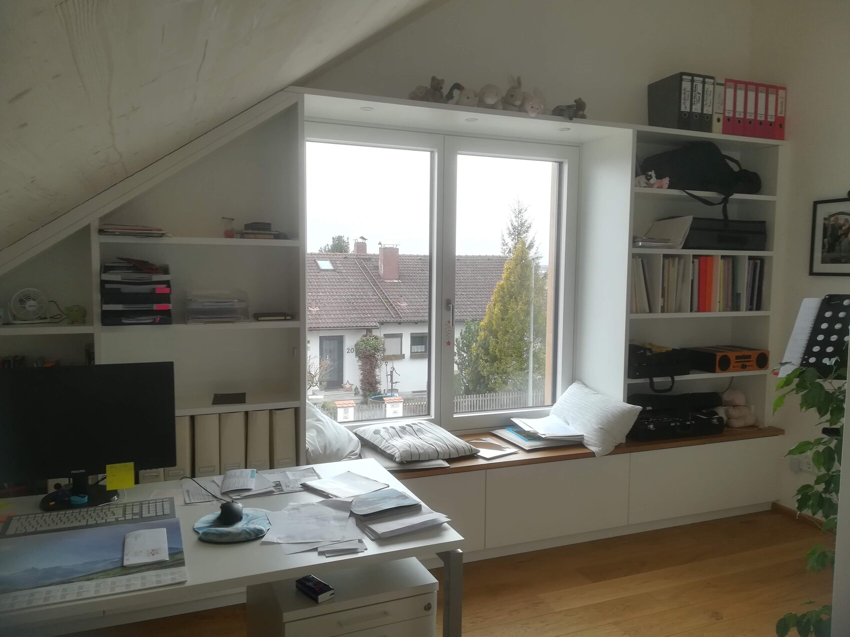 Arbeitszimmer mit weißen Regalen, Fensterbank-Sitzfläche und Schreibtisch voller Unterlagen.