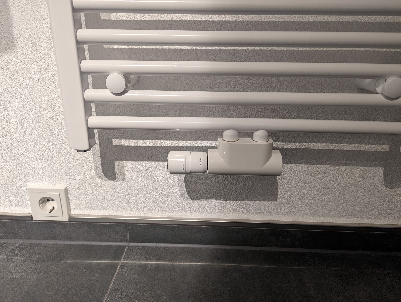 Weißer Handtuchheizkörper mit Thermostatventil, nahe einer Wandsteckdose und dunklen Bodenfliesen.