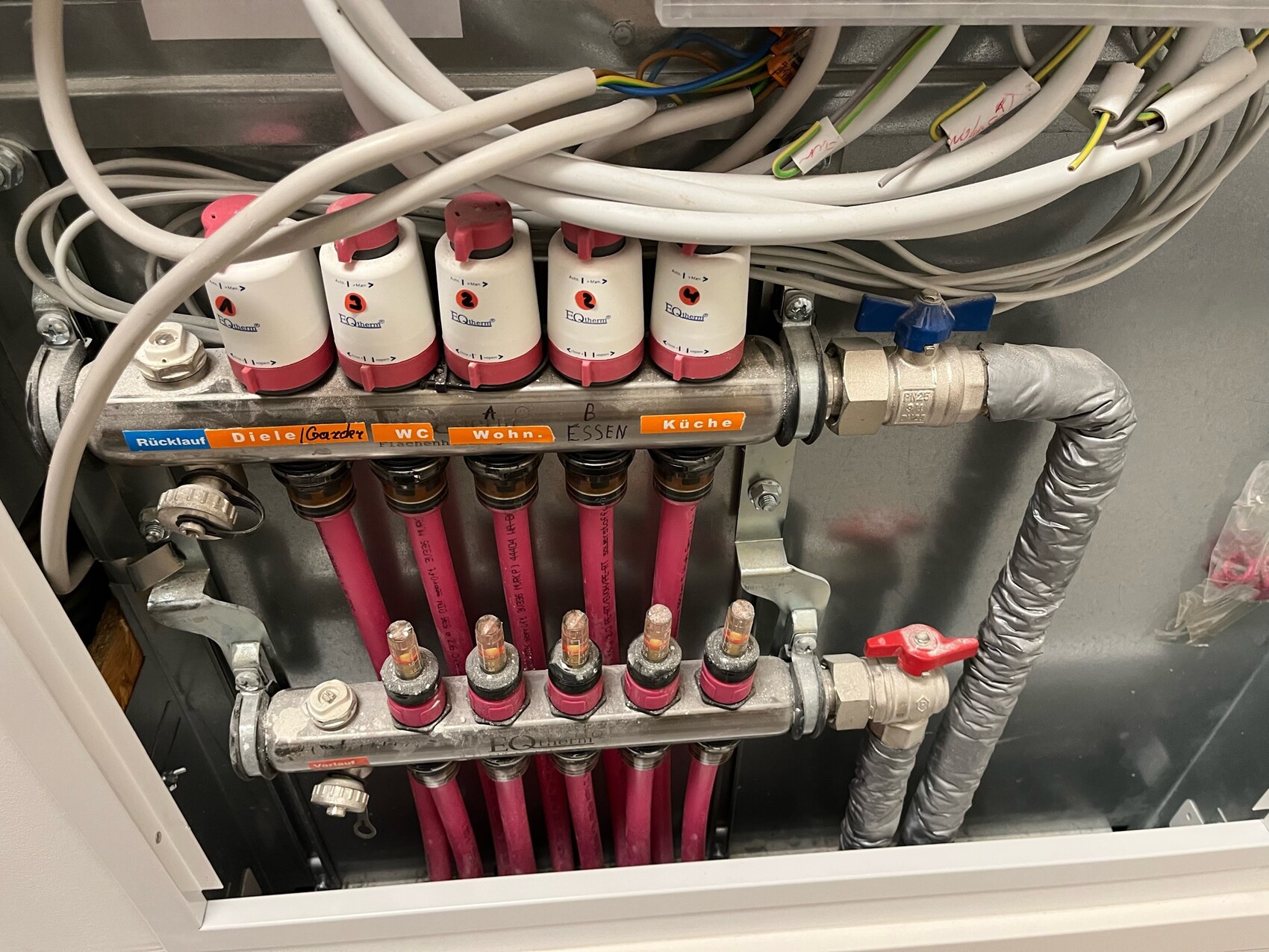 Heizungs-Verteiler mit pinken Rohren, Ventilen und Kabelbündeln im Metallgehäuse.