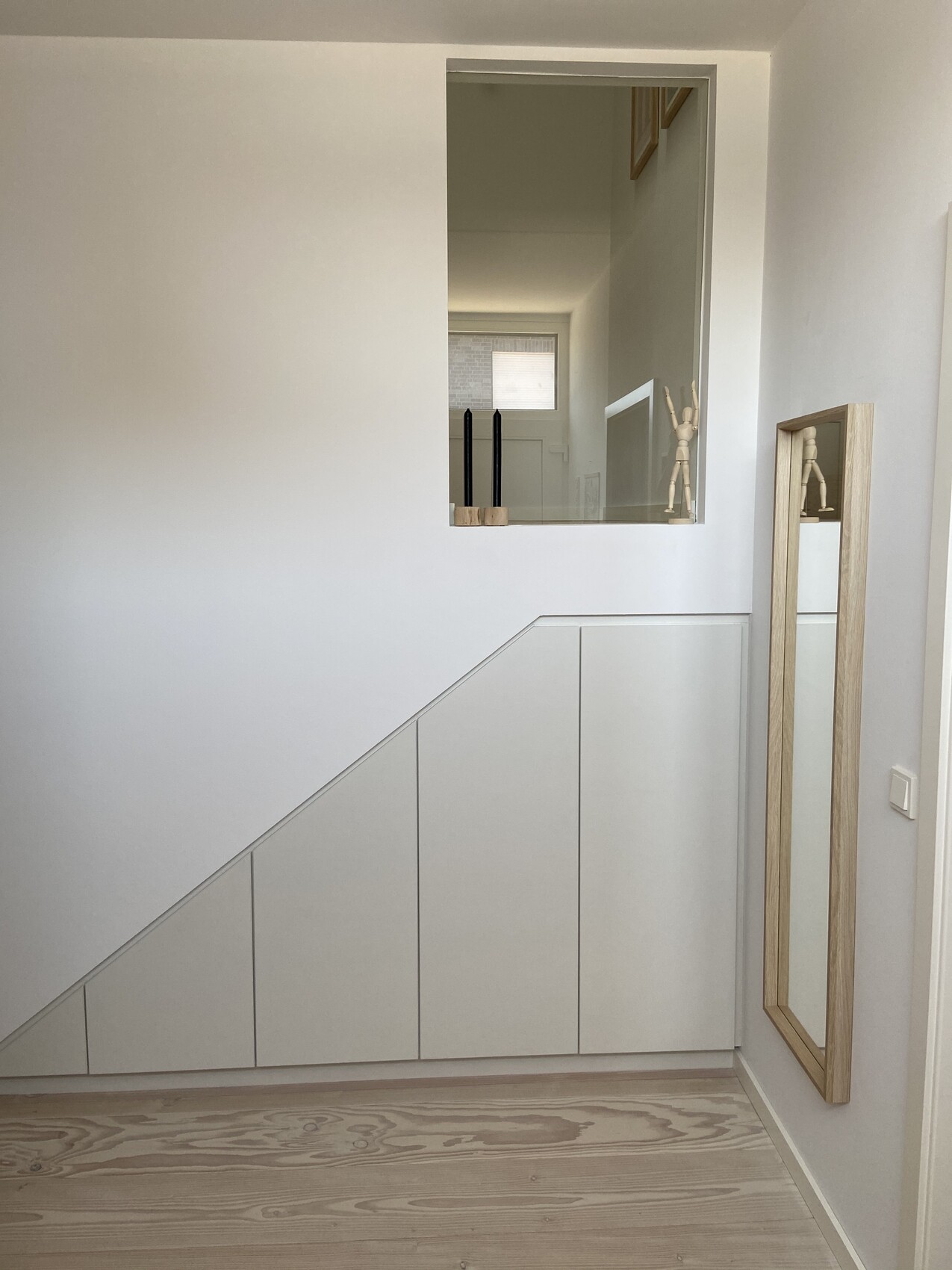 Heller Flur mit weißem Unterschrank unter Treppe; Spiegel rechts, Wandöffnung mit Deko-Objekten.