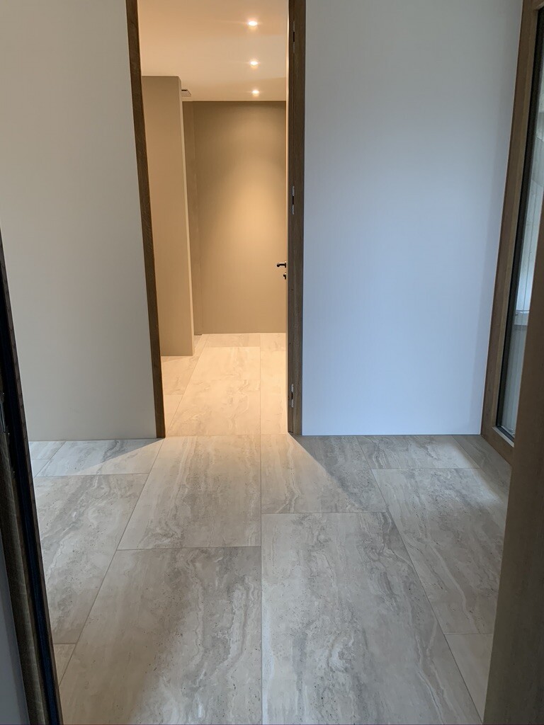 Heller Flur mit offenem Türrahmen, hellgrauer Fliesenboden, beige Wände, Deckenleuchten.