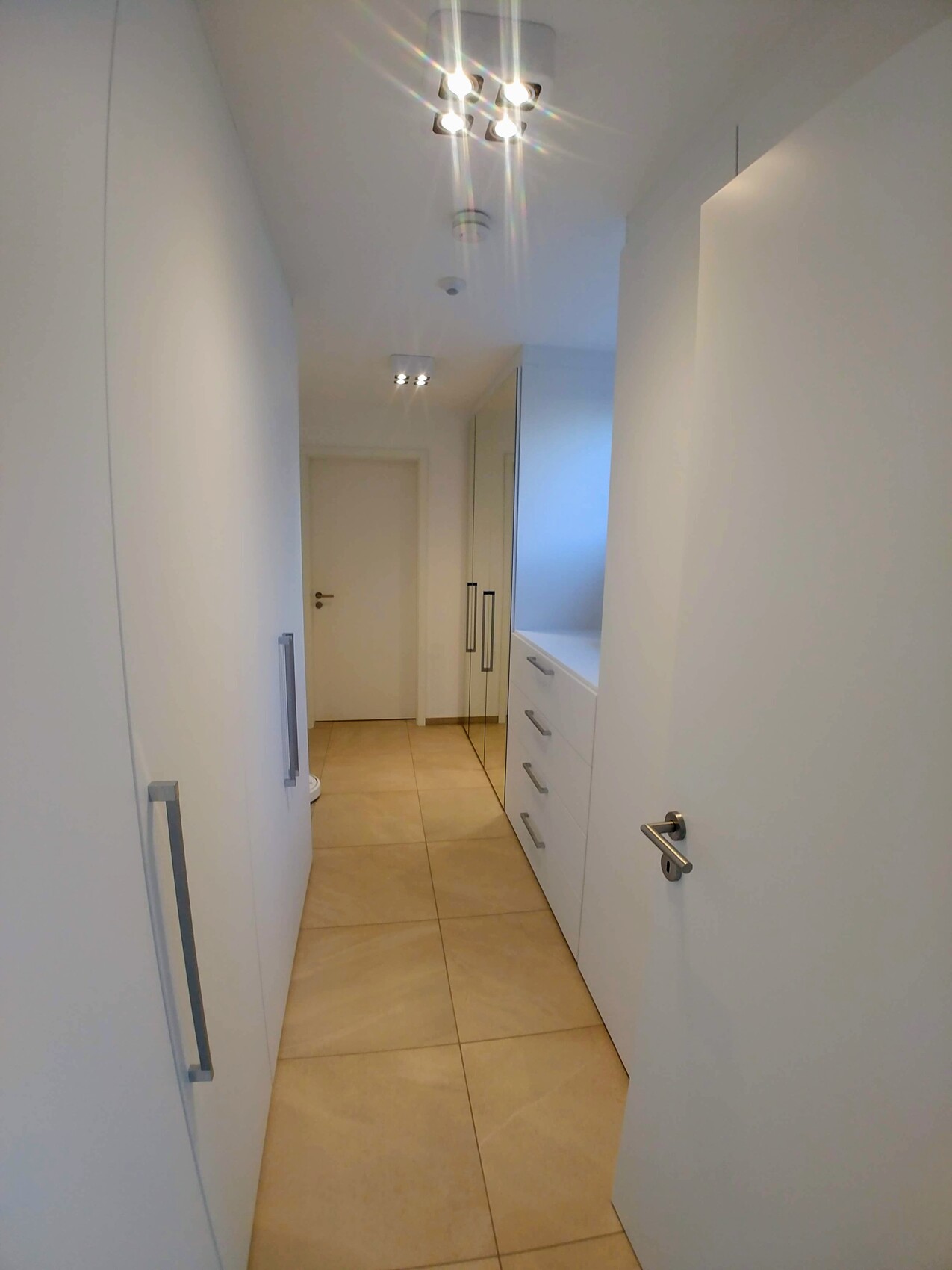 Schmaler Flur mit weißen Schränken links, Schubladen rechts, beige Bodenfliesen, Deckenleuchten.