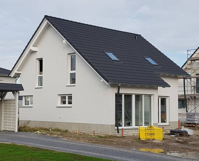 Modernes weißes Einfamilienhaus mit dunklem Satteldach und großen Glasfronten auf einer Baustelle.