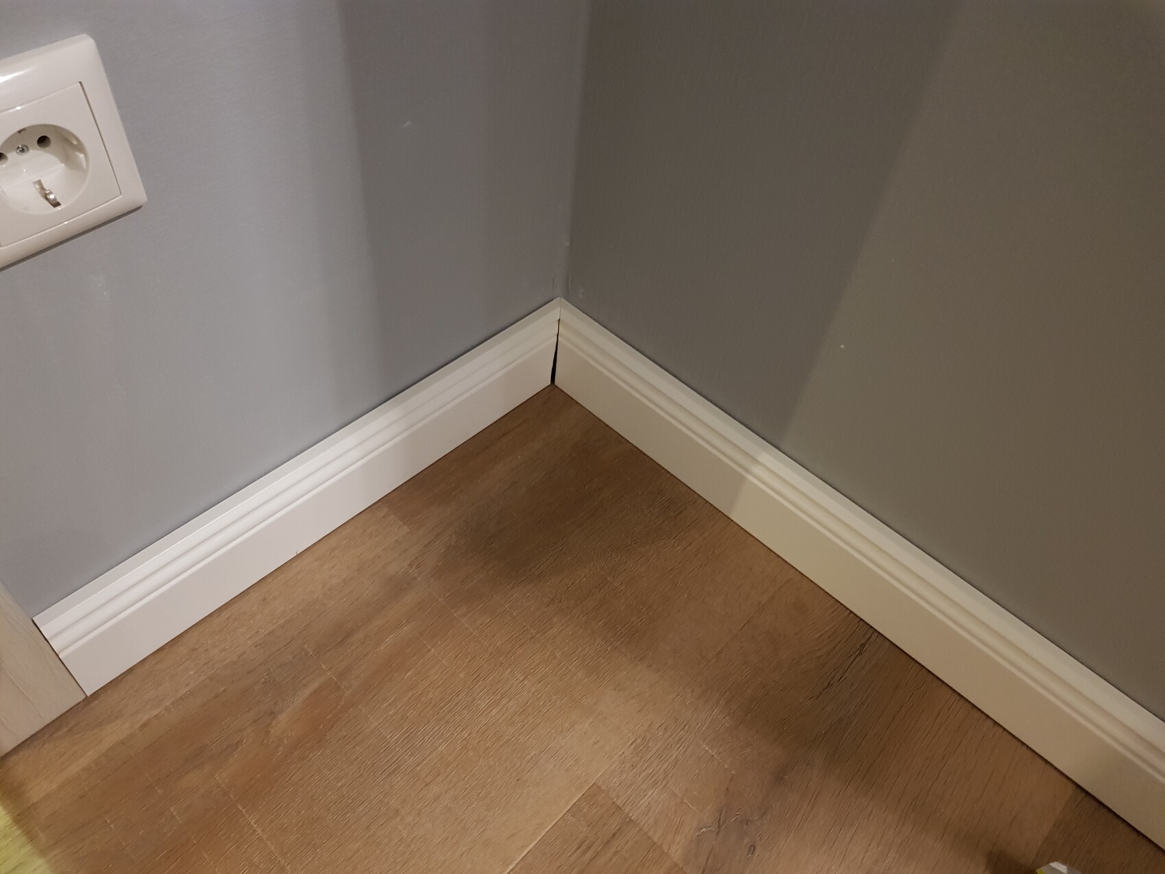 Eckbereich: graue Wände, weiße Sockelleisten, Laminatboden; Steckdose an der linken Wand.