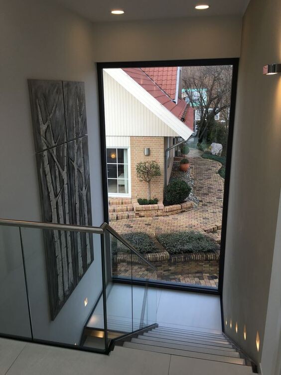 Innenansicht eines Treppenhauses: Glasgeländer, großes Fenster, Blick auf Gartenweg.