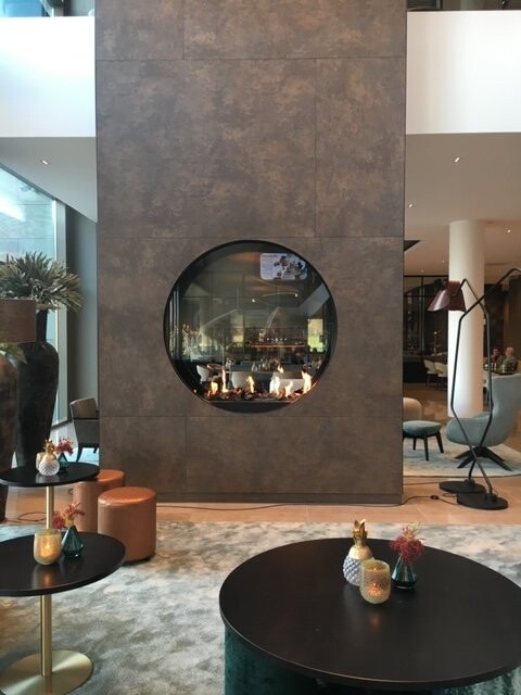 Moderne Hotellounge mit rundem Kaminfenster, Flammen sichtbar, dunkler Wand, Tische und Dekorationen.