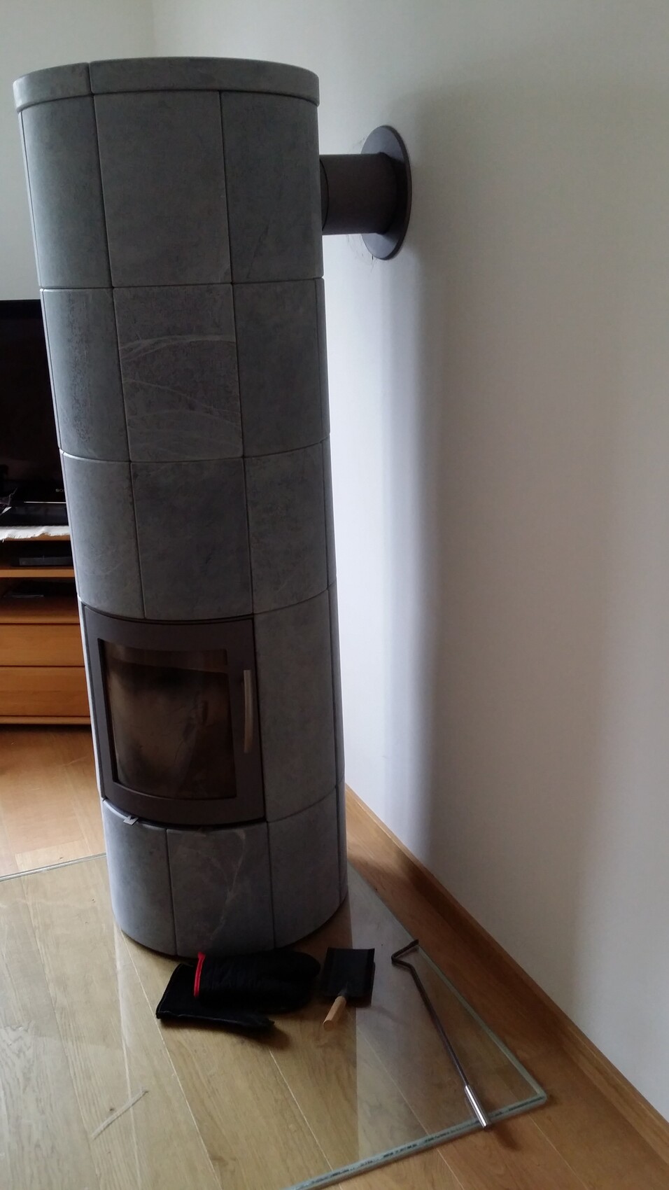Zylindrischer Kaminofen aus grauen Kacheln, Wandmontage, Glastür; Werkzeuge liegen daneben.