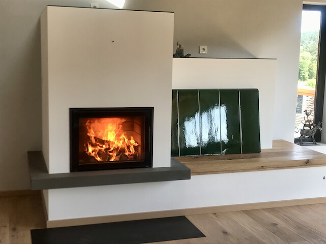 Moderner Kamin mit Glasfront und Flammen; weiße Wände, Holzboden, grüne Fliesen rechts.