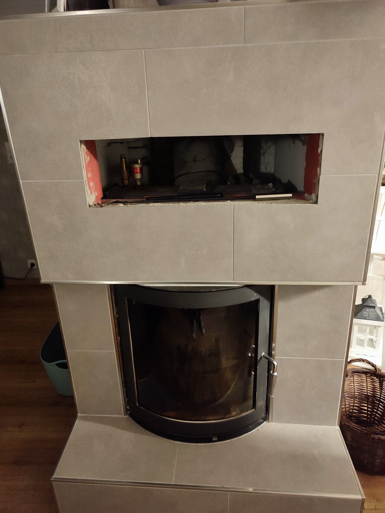 Moderner Kaminofen mit runder Glastür; darüber offenes Fach mit Rohren sichtbar.