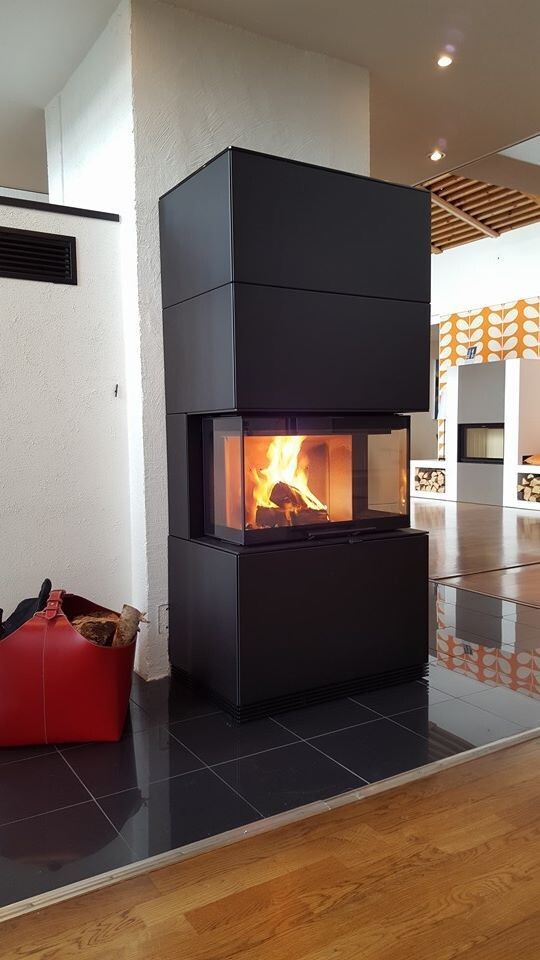Moderner schwarzer Kaminofen mit Glasfront brennt; roter Holzkorb mit Brennholz daneben.