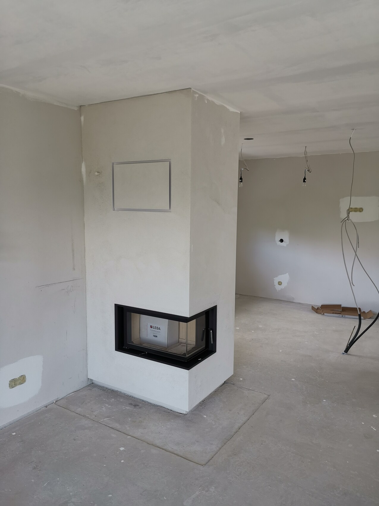 Rohbau-Wohnzimmer mit eingebautem Kaminofen in Säulenstruktur, freiliegende Kabel und unfertige Wände.