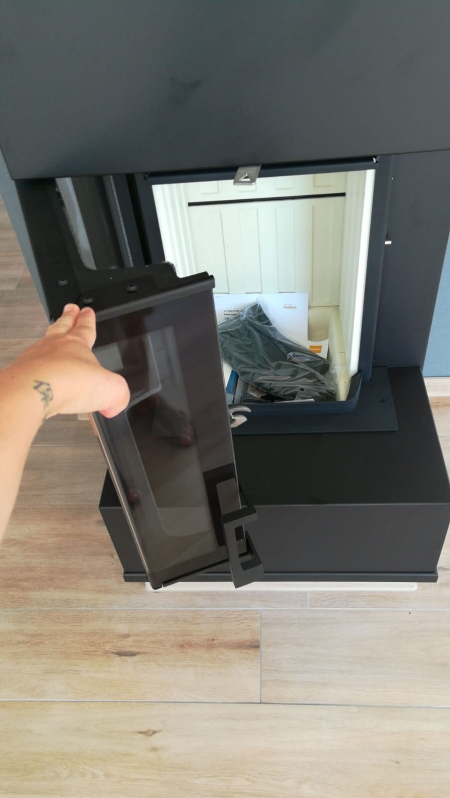 Hand öffnet die Glasfront eines schwarzen Kaminofens; Innenraum sichtbar.