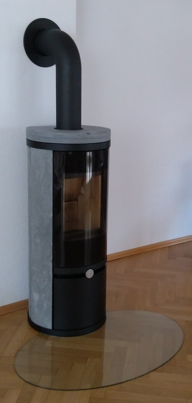 Schwarzer Kaminofen mit Glasfront, gebogenem Abgasrohr, steht auf runder Metallscheibe im Wohnzimmer.