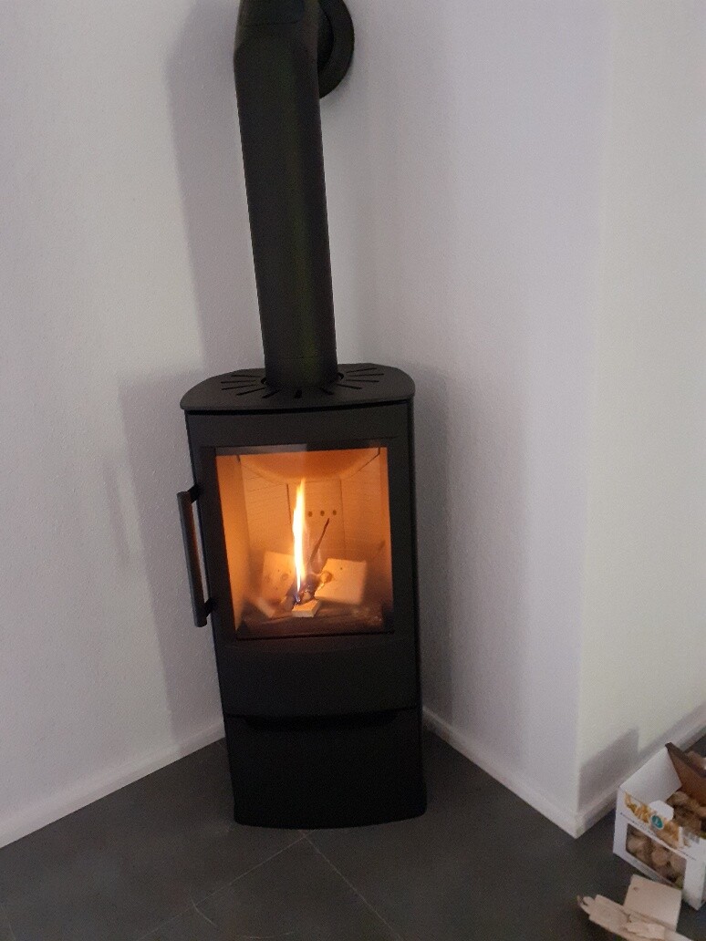 Kleiner schwarzer Kaminofen mit Glastür brennt, Flamme sichtbar, Raumecke mit Holzscheiten.