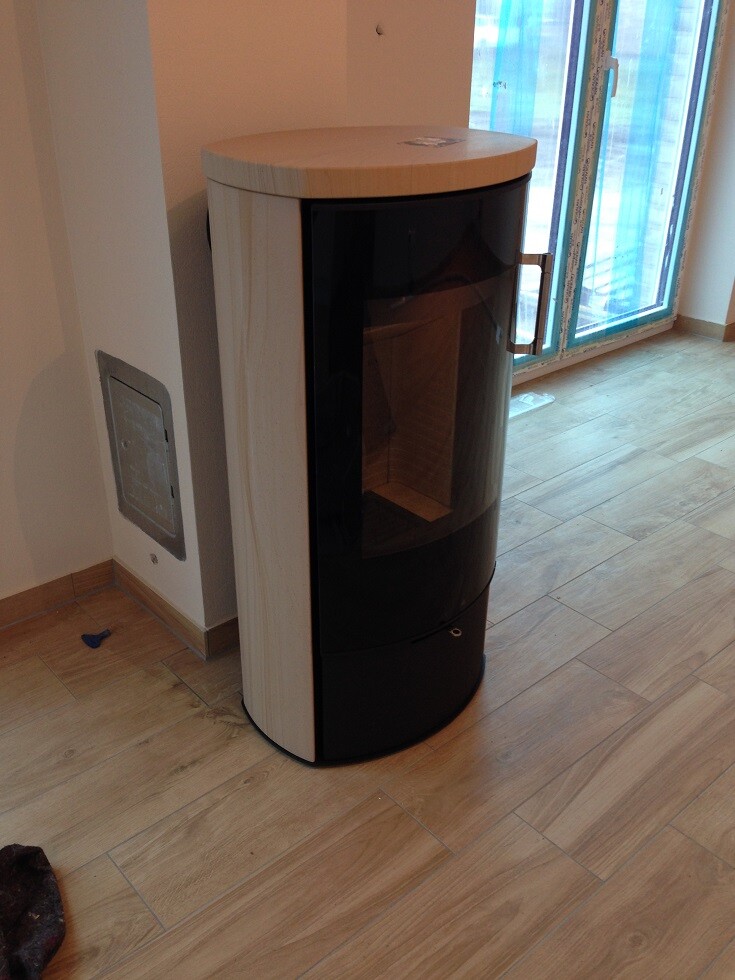 Runder freistehender Kaminofen mit Glasfront und hellem Holzgehäuse im Wohnzimmer neben Terrassentür.