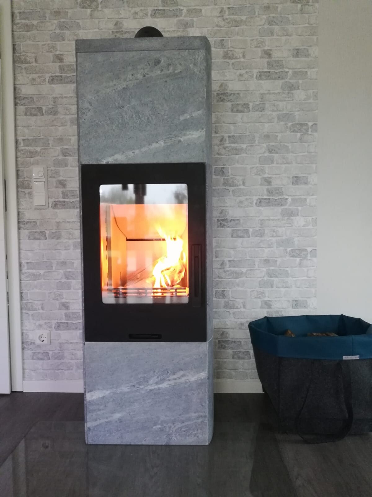 Moderner grauer Kaminofen aus Stein mit brennendem Feuer, daneben ein Holzkorb.