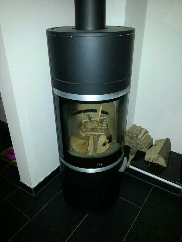 Runder moderner Holzofen mit Glastür; Brennraum mit Holzscheiten, daneben Holzstapel.