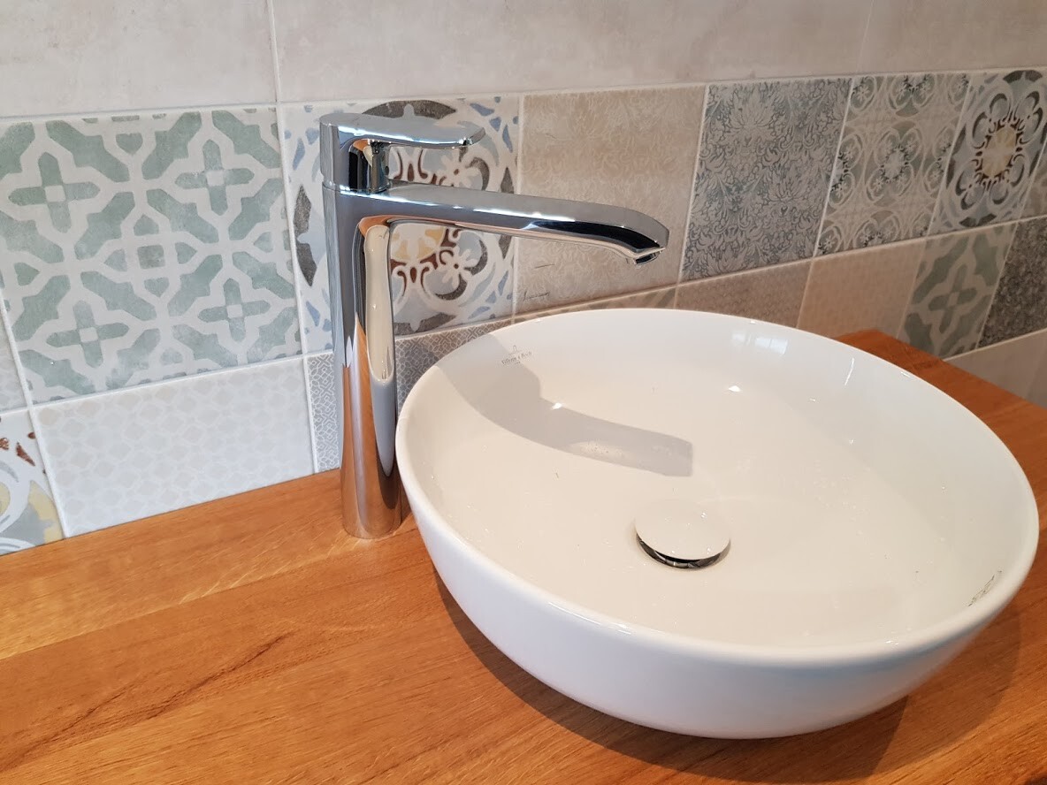 Rundes weißes Becken auf Holzplatte mit Chrom-Armatur vor gemusterten Fliesen.