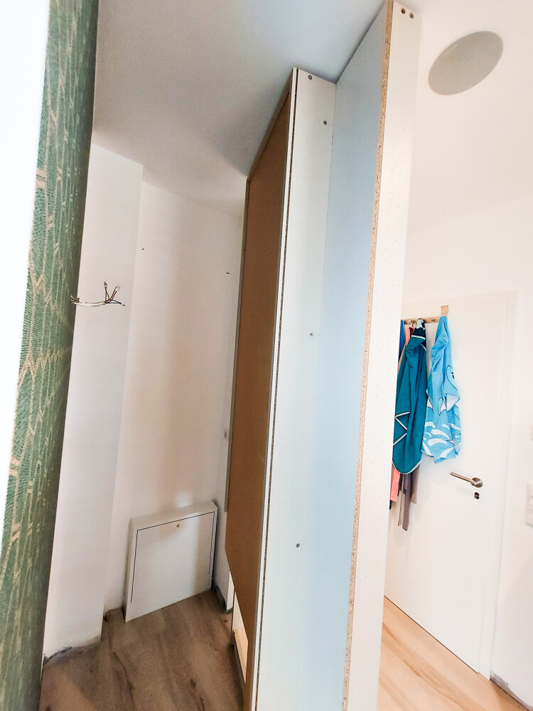 Freistehendes, schmales Möbelteil aus Holzplatten; weiße Wände, Holzboden, blaue Jacken.