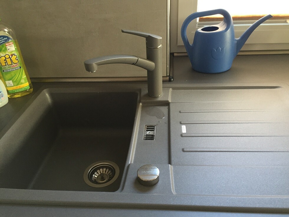 Graue Küchenspüle mit moderner Armatur; blaue Gießkanne am Fenster, Spülmittel links.