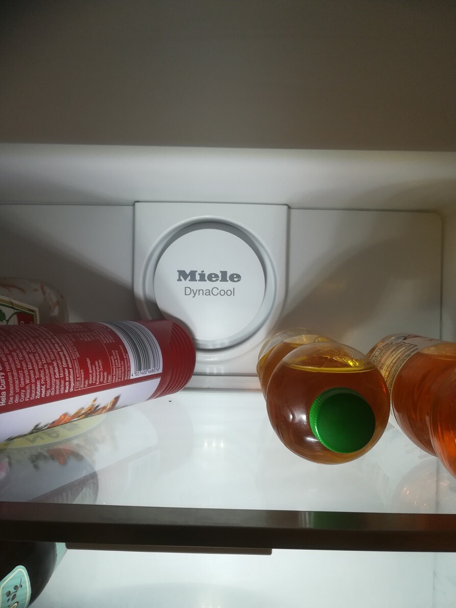 Miele DynaCool-Knopf im Kühlschrank, links rote Dose, rechts orange Flaschen auf Glasboden.