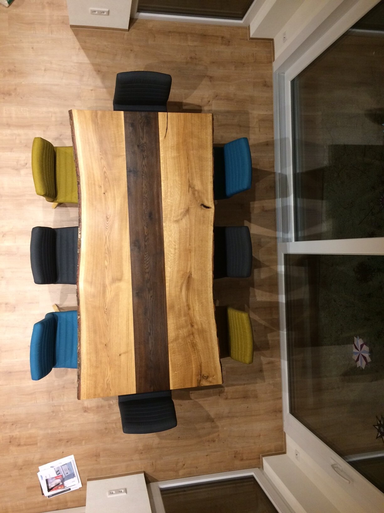 Großes Holztisch mit dunklem Mittelstreifen, bunte Stühle rundum, Holzboden.
