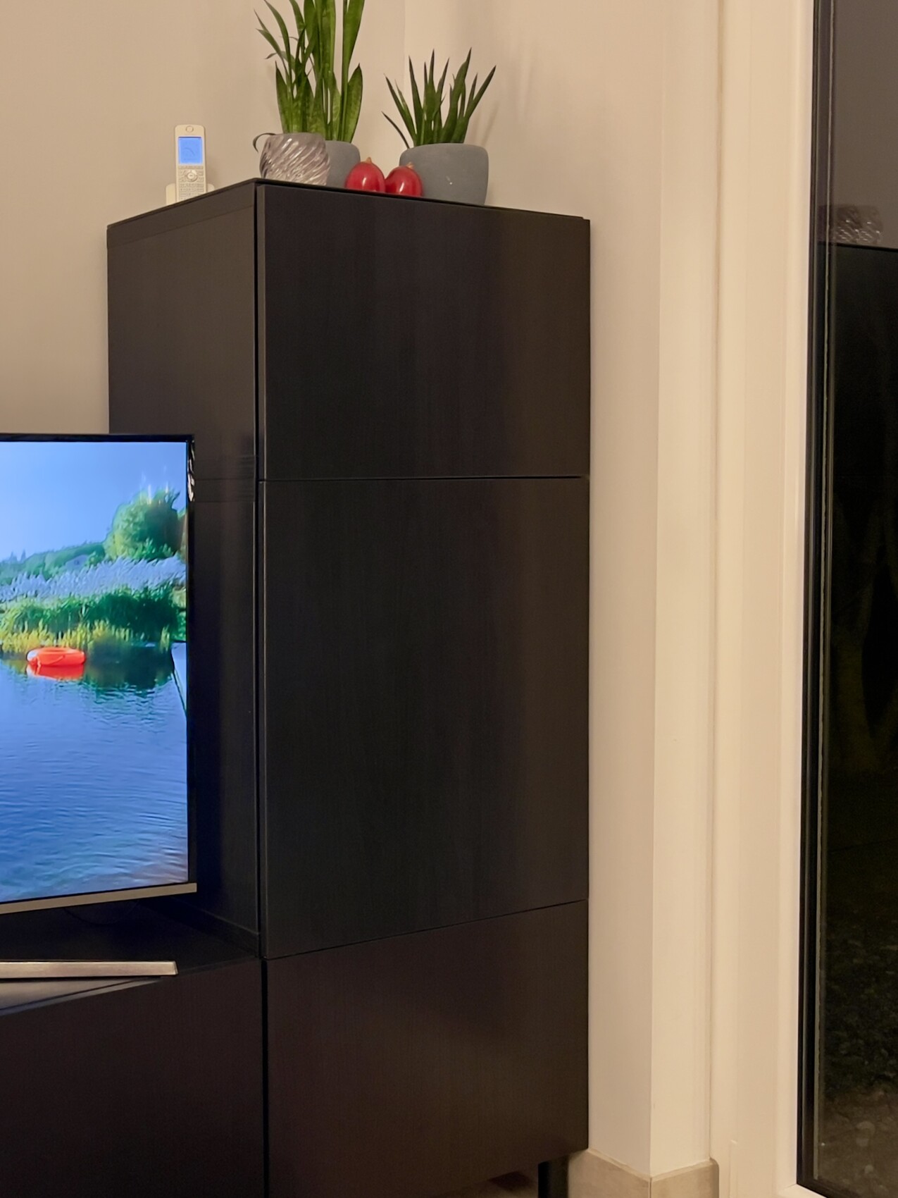 Dunkler TV-Schrank im Wohnzimmer, TV links, oben Pflanzen und rote Äpfel; Fenster rechts.