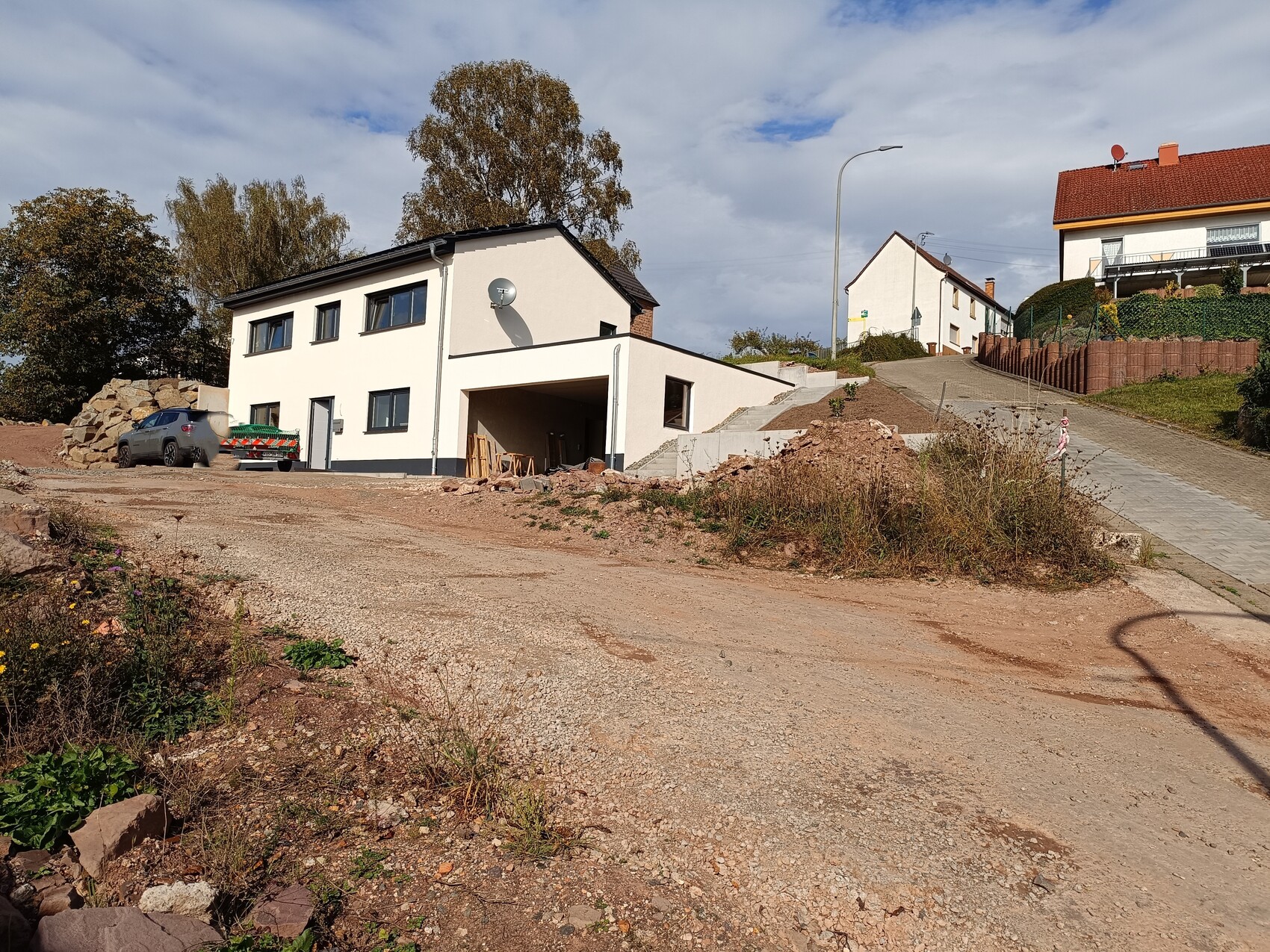 Weiße, zweistöckige Neubau-Villa mit Carport; Bauplatz mit Kies, Steinen und Auto.